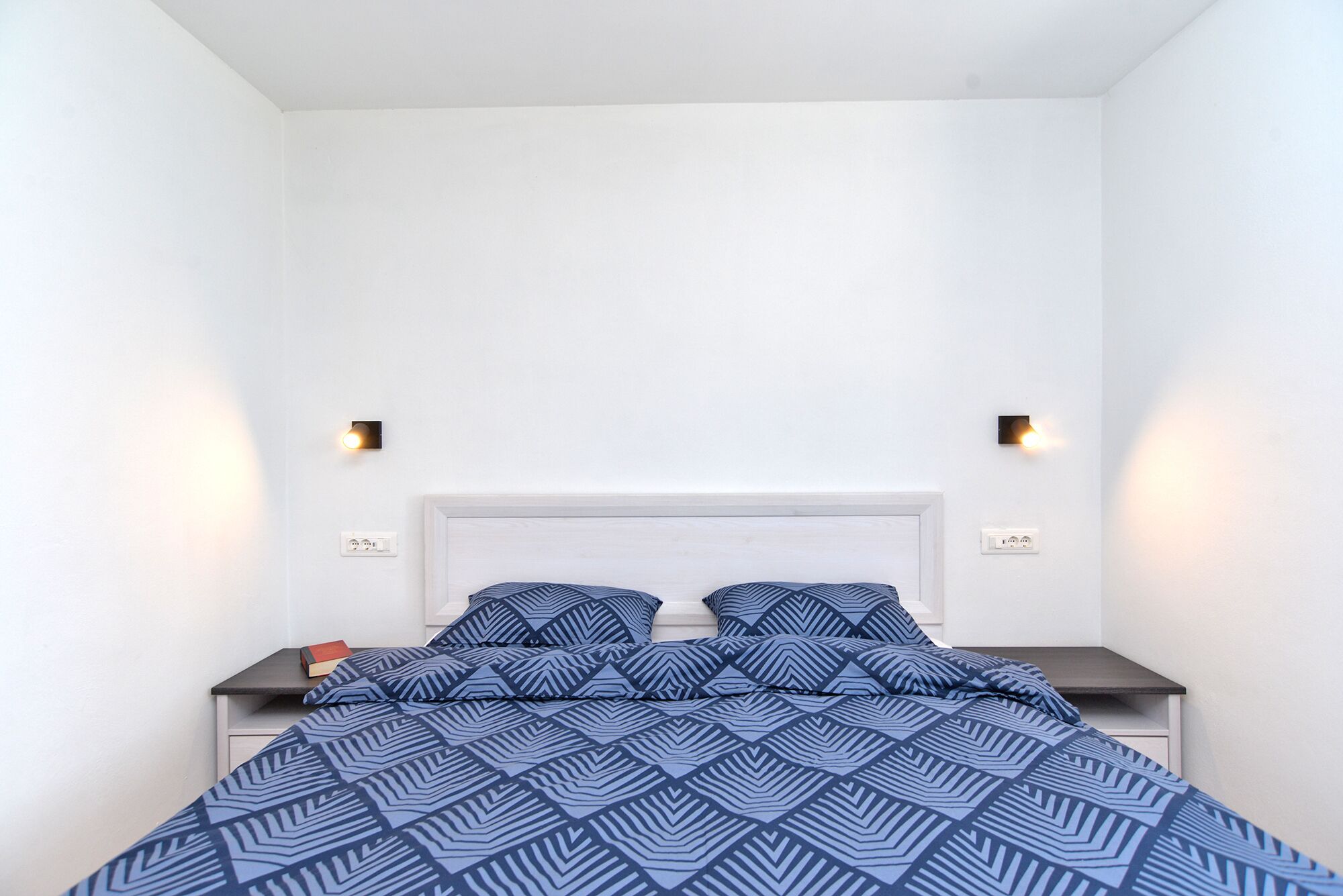 A bedroom with a bed, two bedside tables, and wall-mounted lights.