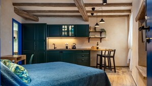 Quarto acolhedor com cama, mesa pequena, cadeira, área de cozinha com armários e pia, e teto de madeira.