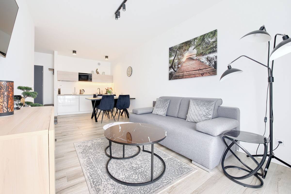 Ein modernes Wohnzimmer mit einer grauen Couch, einem Couchtisch und einer Stehlampe. Im Hintergrund ist eine Küche mit Essbereich zu sehen.