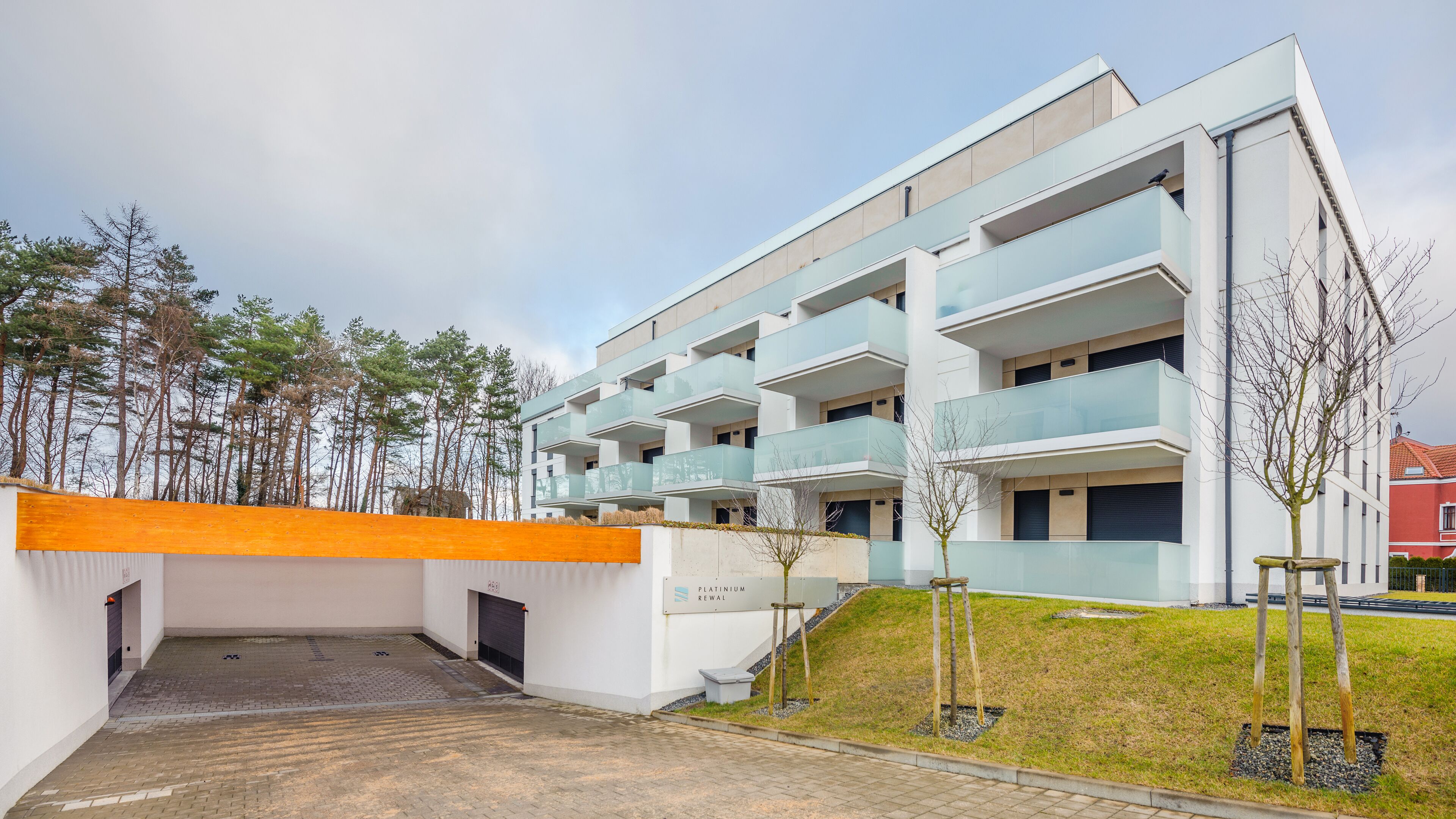 Un edifici residencial modern de diverses plantes, amb balcons, un accés tancat per reixa i una petita zona enjardinada amb arbres joves.