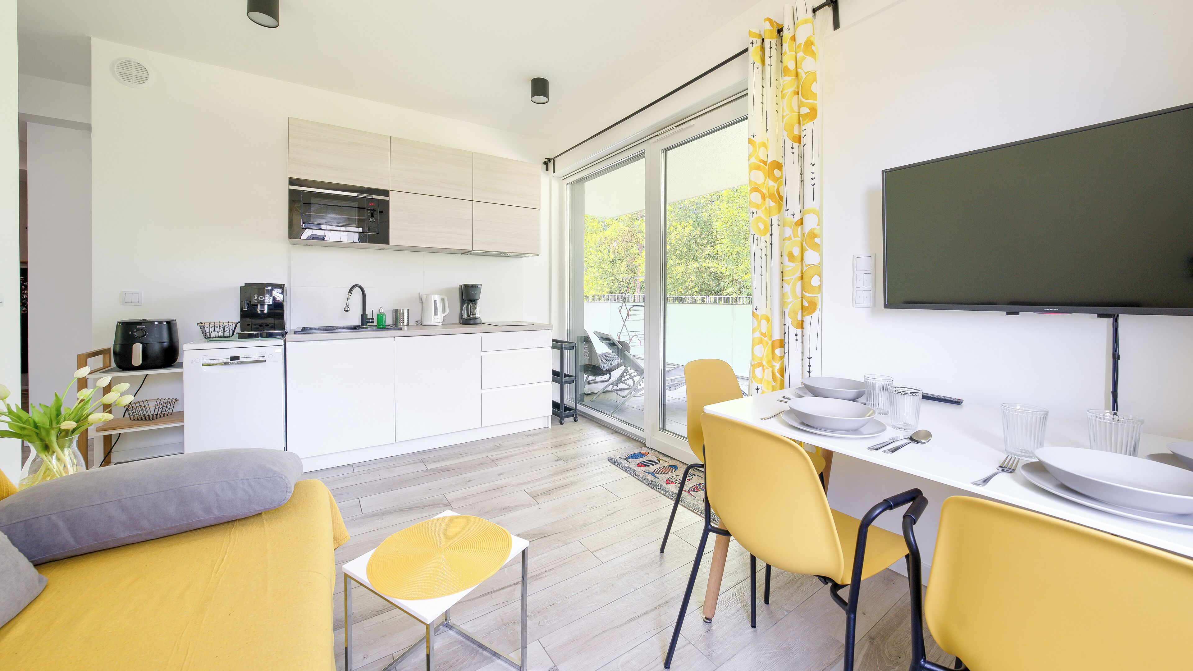 A modern kitchen with white cabinets, a built-in oven, and a dining area with yellow chairs and a white table.