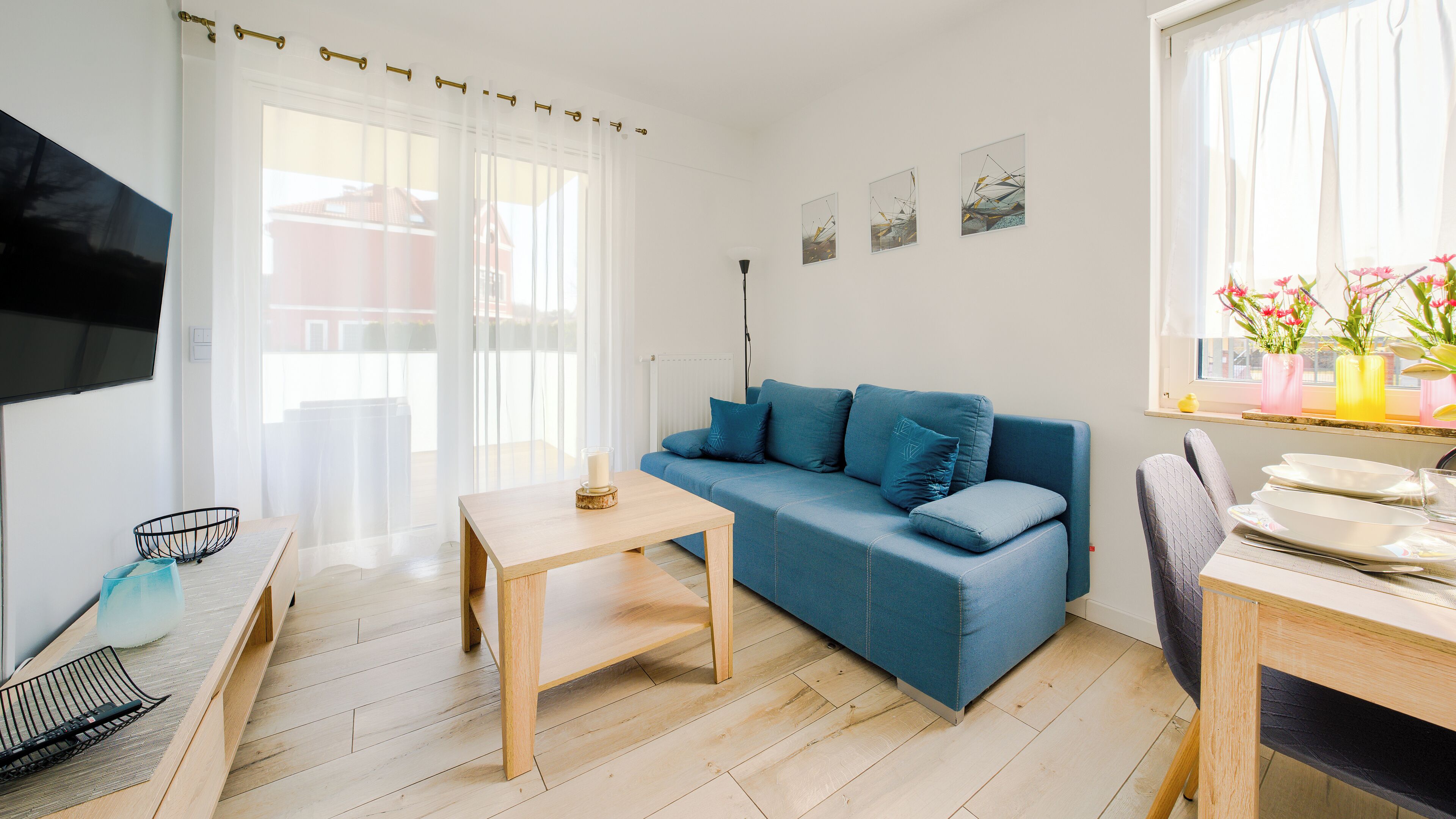 A living room with a blue sofa, a wooden coffee table, a TV mounted on the wall, and a dining area with a table and chairs.