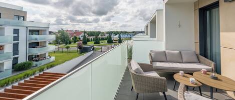 A balcony with wicker furniture, a glass railing, and a view of neighboring buildings and greenery.