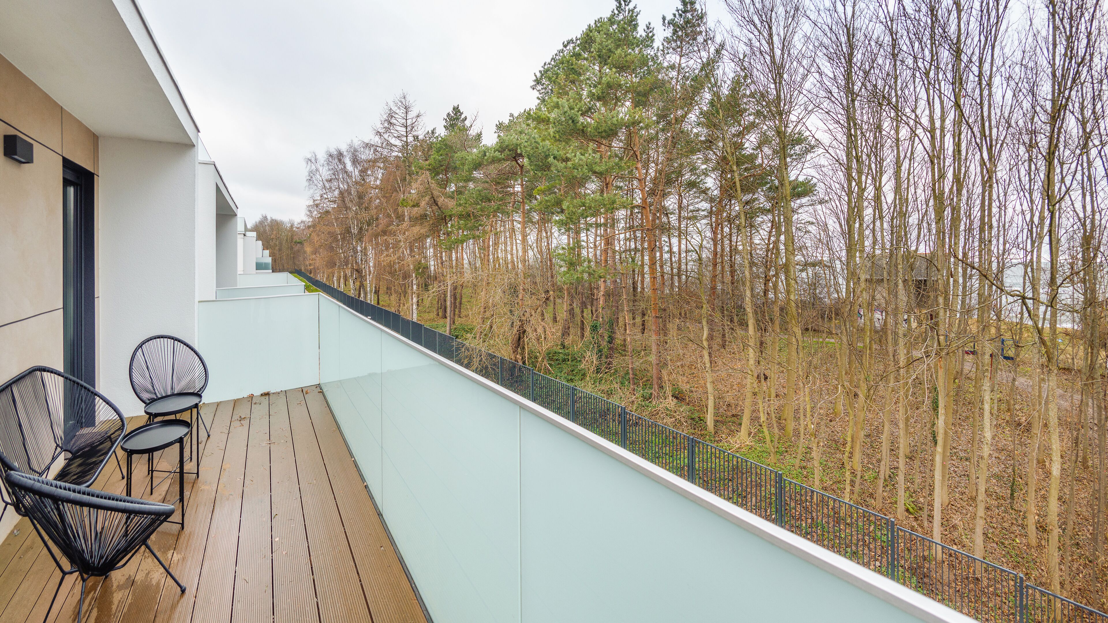 A balcony with a glass railing, wooden floor, and two black chairs.