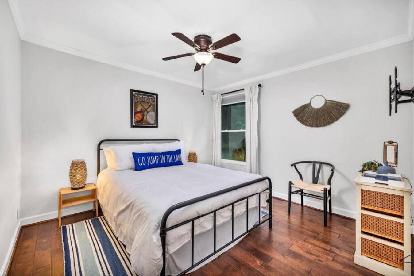 Un dormitorio con cama, ventilador de techo, ventana con cortinas, una silla, mesita de noche y ventilador de pared.