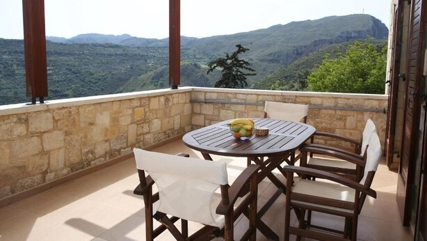 A balcony with a stone wall, wooden chairs, a round table, and a bowl of fruit. - Vasiiliki's Residence-Breathtaking View (Kissamos)