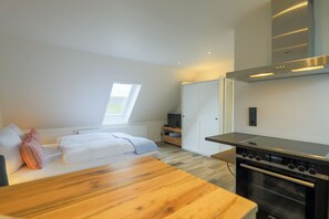 A modern bedroom with a wooden bed, a white wardrobe, a TV, and a kitchen area with a stove. - Apartment (Westerland)