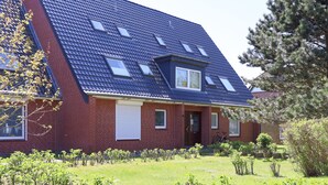 Une maison à deux étages, dotée d’un toit en pente, d’une façade en briques et d’un petit jardin devant, où diverses plantes sont cultivées.