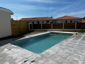 A rectangular swimming pool with a tiled deck, surrounded by a wooden fence and a building with a red roof.