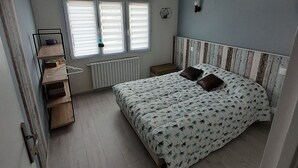 Un dormitorio con cama, radiador, ventana con persianas, estante con libros y lámpara fijada a la pared.