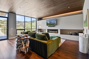 A modern living room with a green sofa, a fireplace, a mounted TV, and a large window overlooking a mountainous landscape.