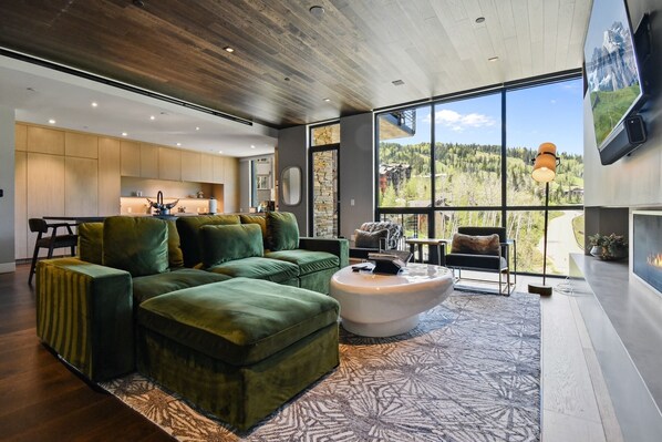 A modern living room with a green sofa set, a white coffee table, and a large window offering a mountain view.