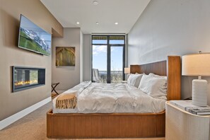 A modern hotel room with a large bed, a fireplace, a television, and a view of the outdoors.