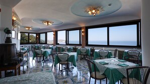 A restaurant with tables set for dining, large windows offering a view of the sea, and decorative ceiling lights. - Hotel Residence Parco Mare Monte (Casamicciola Terme)