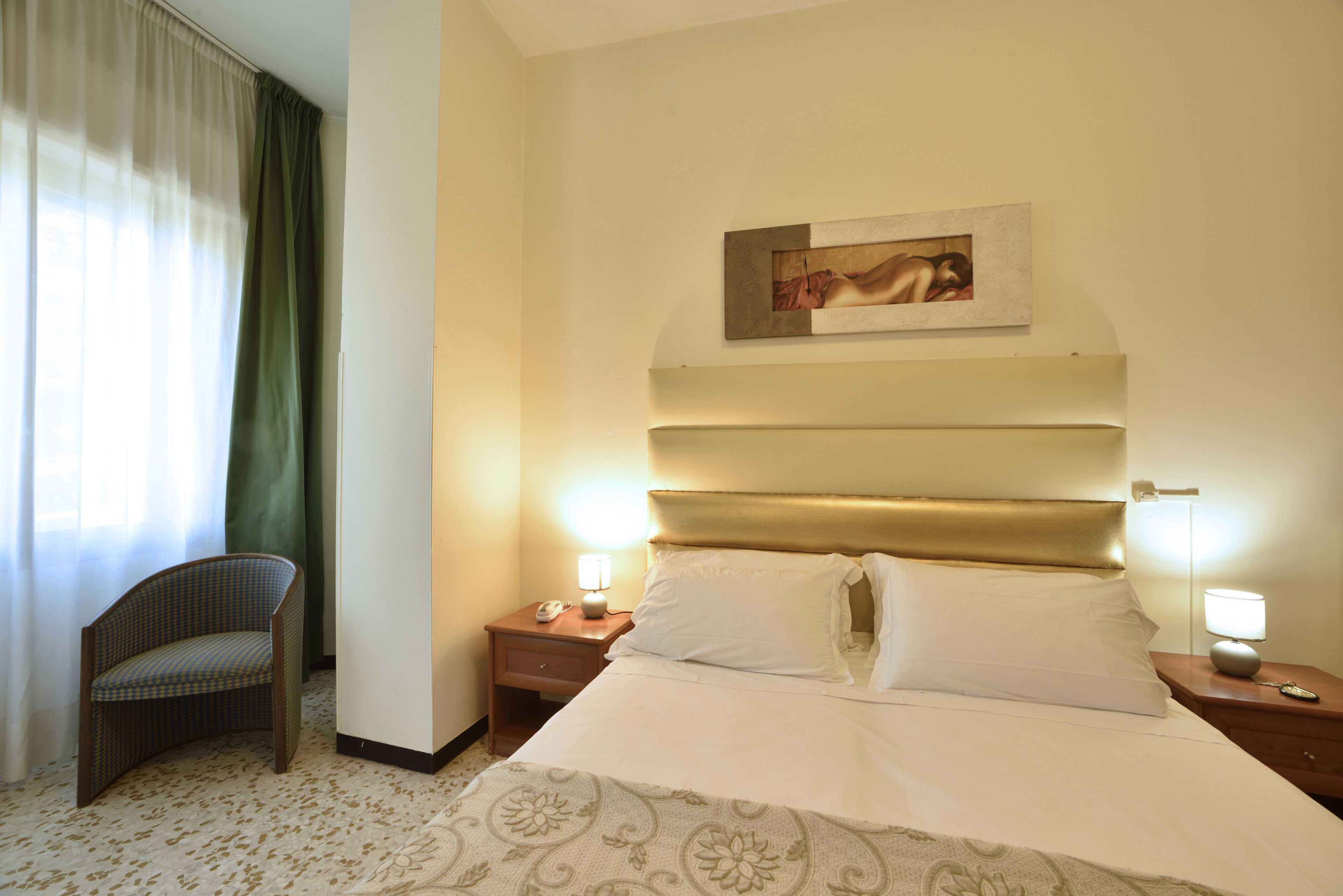 A hotel room with a bed, a chair, two bedside tables with lamps, a painting on the wall, and a window with curtains.