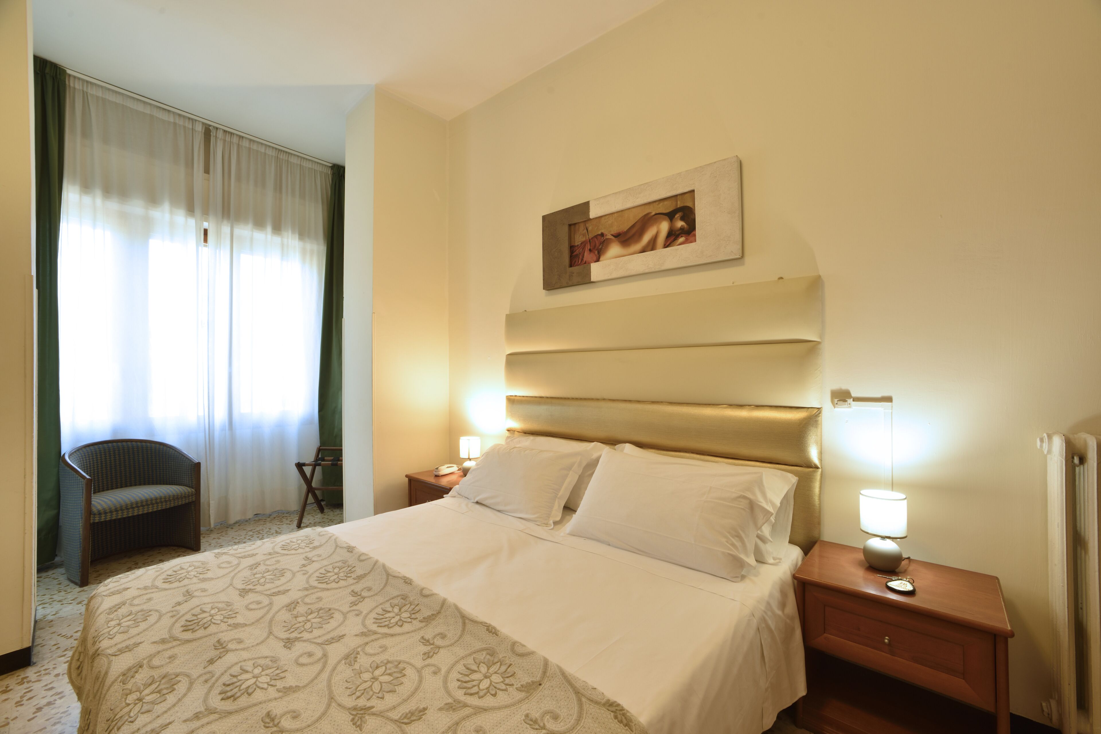 A hotel room with a large bed, a chair, a nightstand with a lamp, and a painting on the wall.