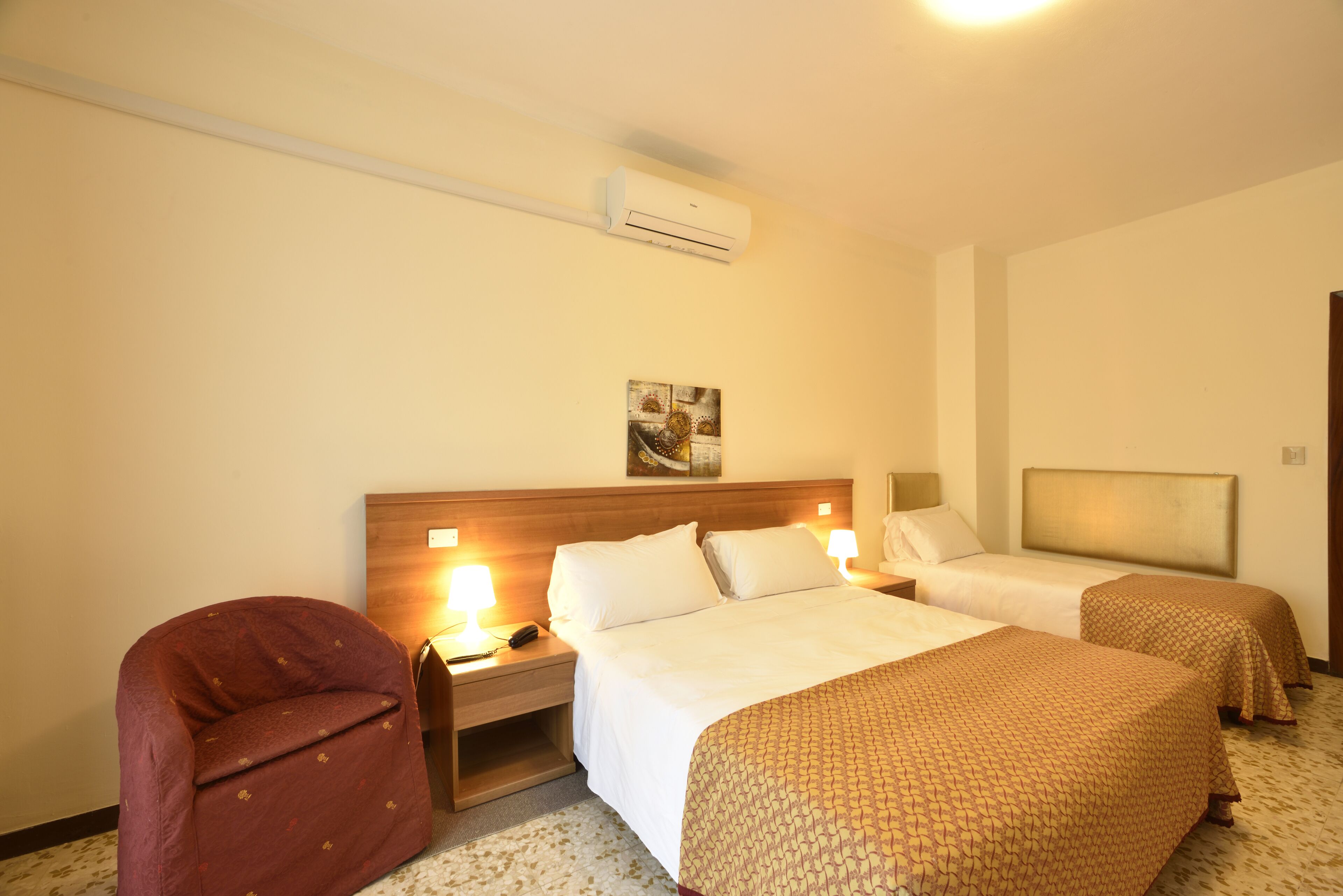 Ein Hotelzimmer mit zwei Betten, einem Nachttisch mit Lampe, einem Sessel und einer Klimaanlage.