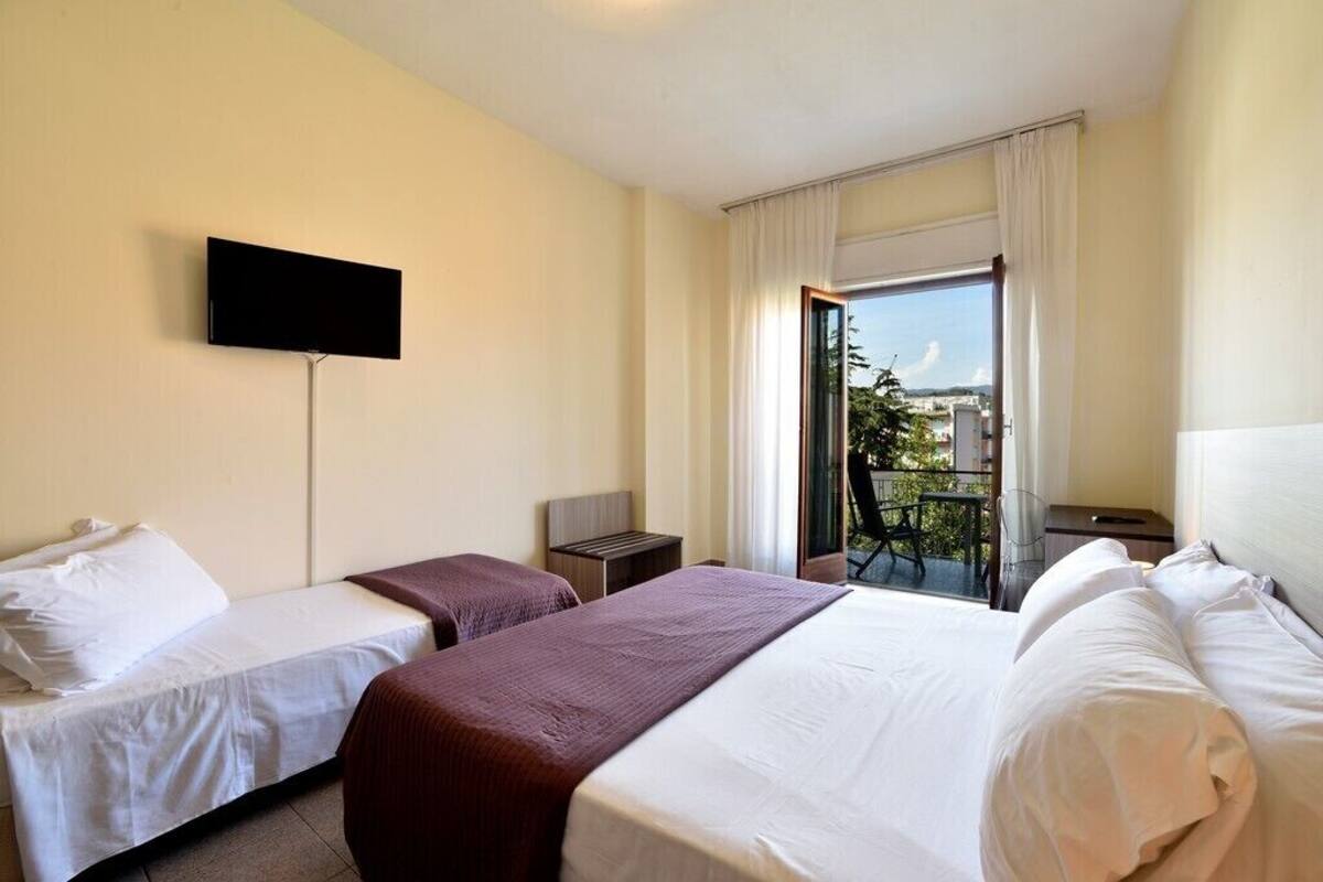 A hotel room with two beds, a TV mounted on the wall, a small table, and a balcony with a view of the city.