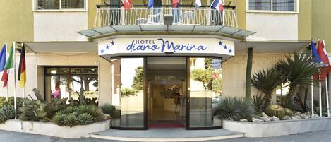 A hotel named "Hotel d'ano Marina" with a balcony, flags, and a red carpet entrance.