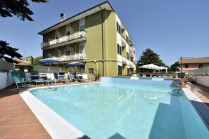 Una piscina con sillas de descanso y sombrillas, rodeada por un edificio de varios pisos con balcones.