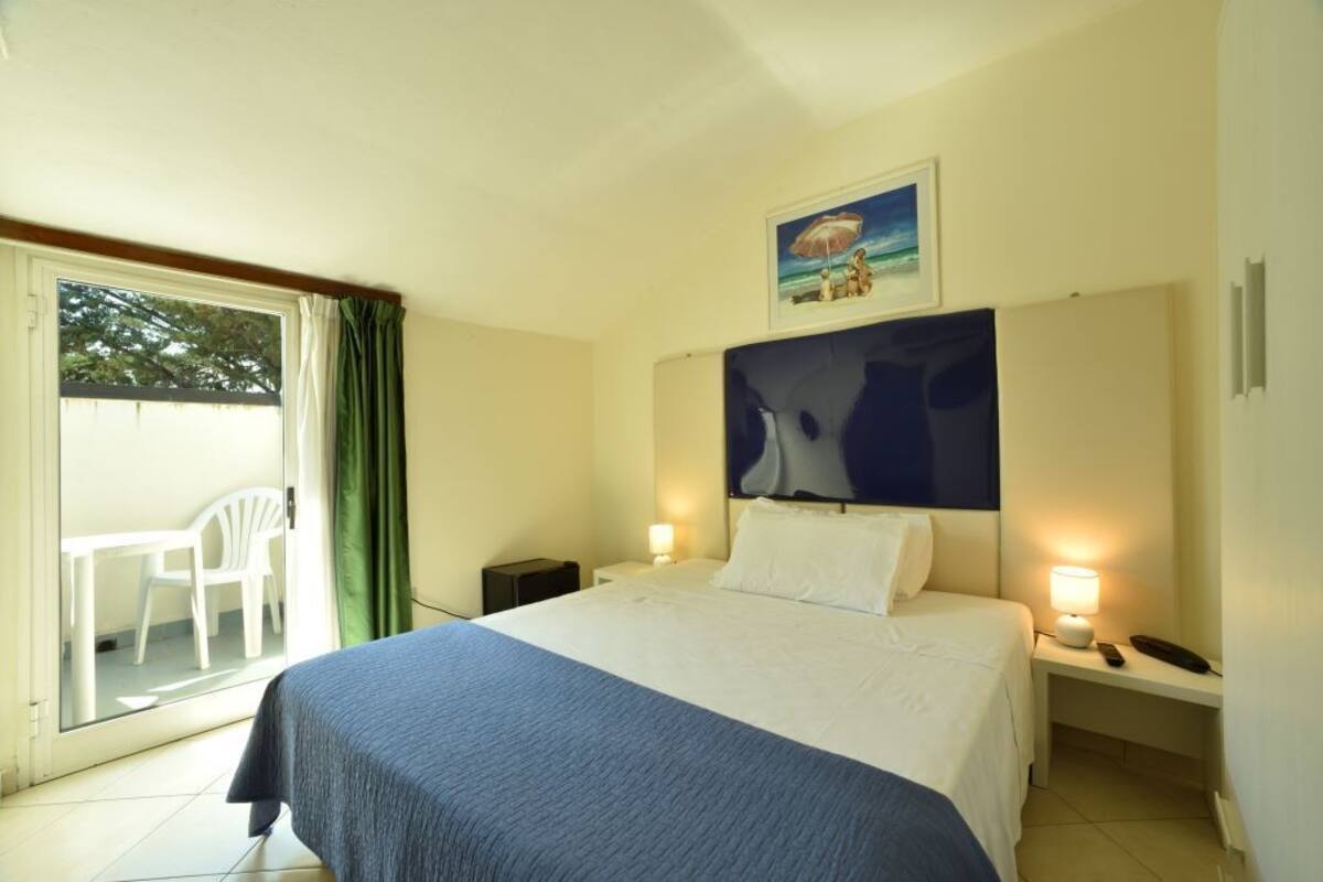 A hotel room with a bed, a nightstand with a lamp, a painting above the bed, and a view of the outdoors through a sliding door.