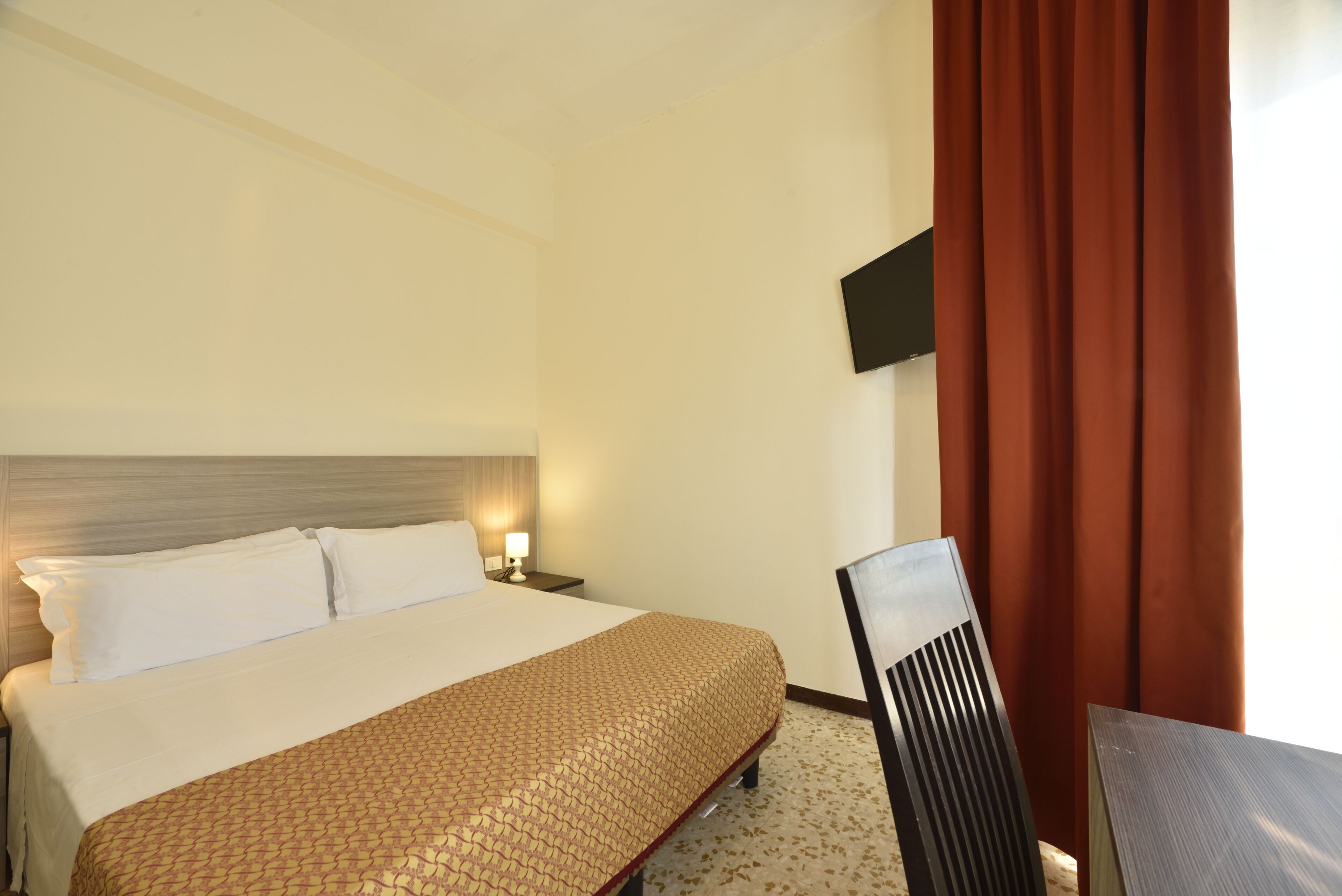 A hotel room with a bed, a nightstand with a lamp, a TV mounted on the wall, and a chair.