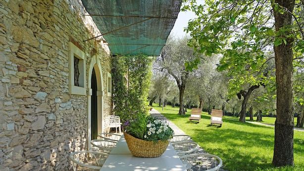 A stone building with a covered outdoor seating area featuring a table with chairs and a large woven basket with flowers. - Villa Gran Paradiso (Toscolano-Maderno)