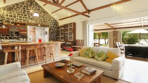 A spacious living area with a stone wall, wooden beams, a kitchen with a stone backsplash, and a large window overlooking a garden. - Villa (Mahebourg)