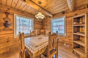 A wooden cabin interior with a dining table, chairs, a chandelier, and a bookshelf.