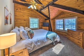 A cozy bedroom with a wooden ceiling, a bed with pillows, a TV, and two windows.