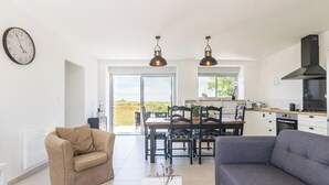 A modern living room with a sofa, a chair, a dining table with chairs, a kitchen area, and a large window.