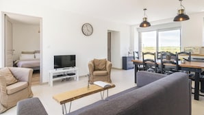 A modern living room with a sofa, a TV, a dining area with chairs, and a large window offering a view.