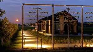 Et overdækket udendørs område med en pergola, en sti til en bygning, og en solnedgang i baggrunden.
