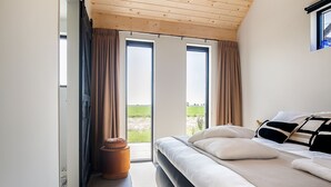 Et soveværelse med en stor seng, to vinduer med gardiner, udsigt til det fri og et træloft.