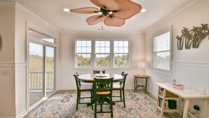 A dining area with a round table, four chairs, a ceiling fan, and a view through large windows.