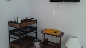 A room with a TV mounted on the wall, a wooden shelf unit, a small table with a tray, a lamp, and a trash can.