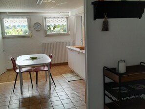 A small, bright kitchen with a dining table and chairs, two windows with heart decorations, a clock, and a radiator.