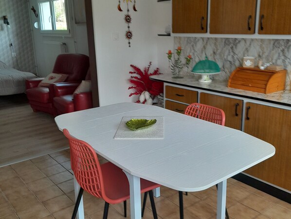 A dining area with a white table, red chairs, a green decorative plate, and a kitchen in the background.