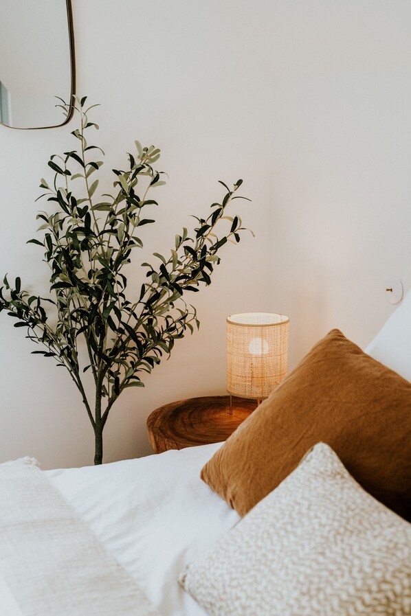A neatly made bed with white linens and two pillows, a small wooden side table with a woven lamp, and a plant with green leaves.