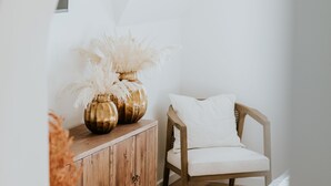A wooden cabinet with two gold vases containing white flowers, a wooden chair with a white cushion, and a white bed in a room with wooden flooring and white walls. - La Brise (Roscoff)