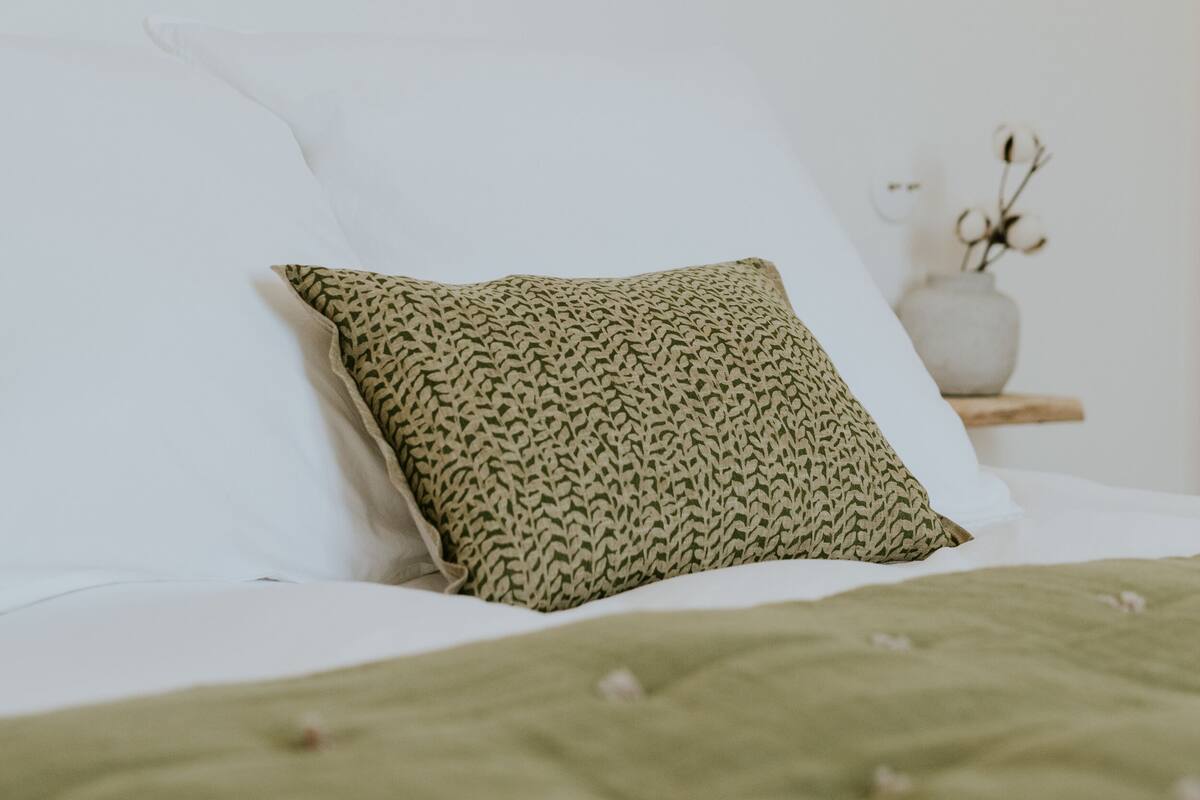 Una cama bien hecha con un edredón verde y almohadas estampadas, una mesita de noche con un jarrón de flores secas, y una lámpara de pared.