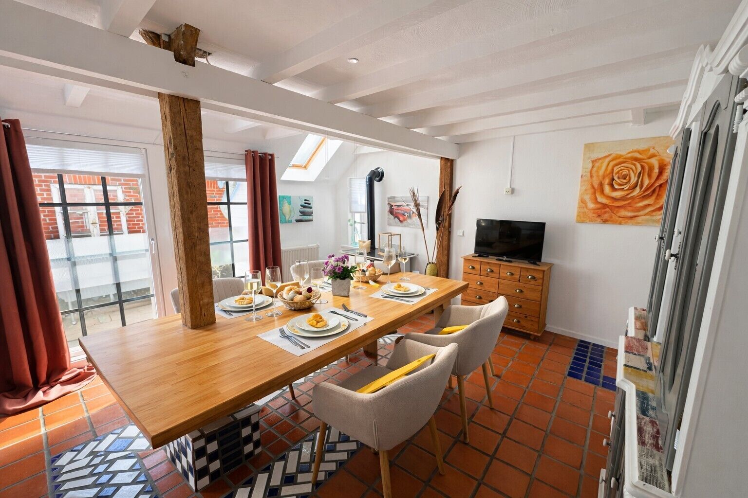 Un comedor con una mesa de madera para cuatro, sillas blancas, un televisor sobre un mueble de madera y un ventanal con cortinas.