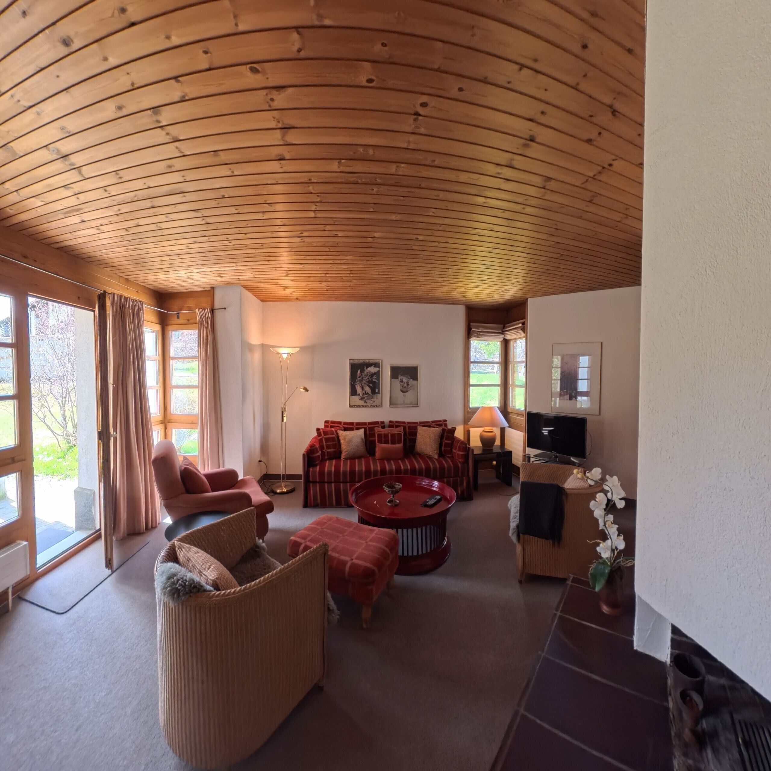 Uma sala de estar acolhedora com teto de madeira curvada, um sofá vermelho, uma cadeira de vime, uma mesa de centro vermelha e uma televisão.