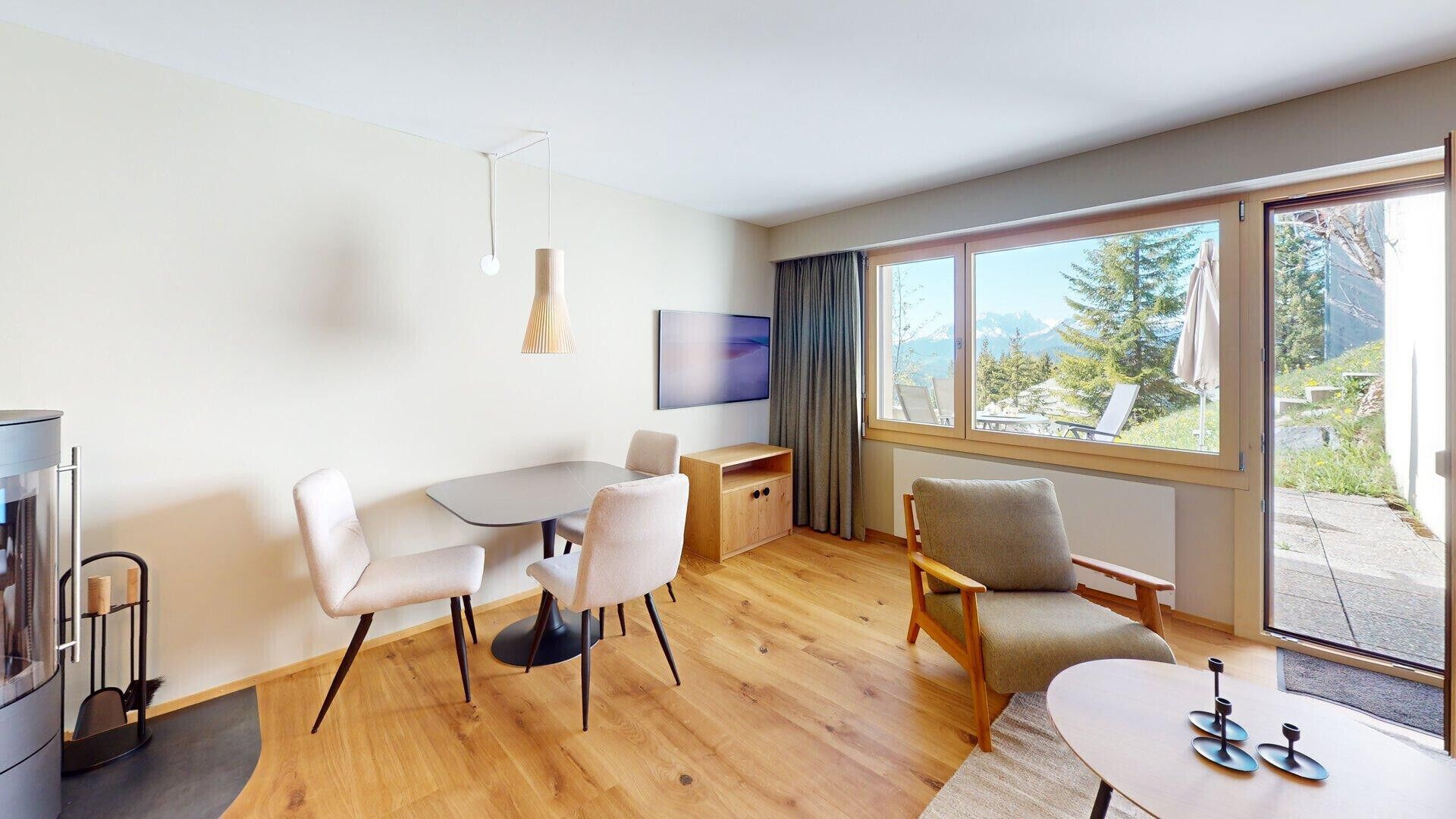 Una sala de estar moderna con comedor, televisor y un amplio ventanal con vistas a árboles y montañas.