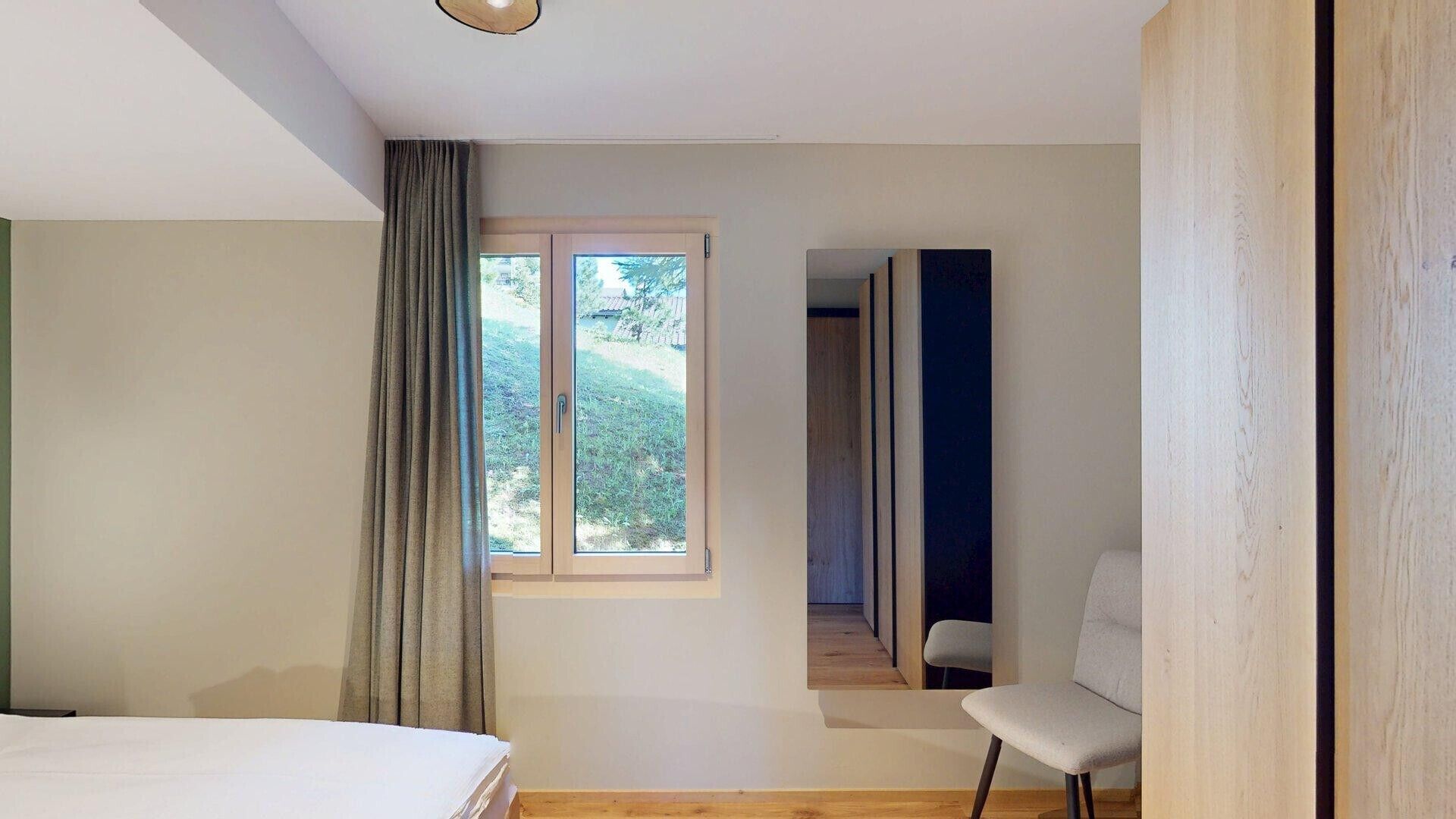 A bedroom with a bed, a window with curtains, a chair, and a view of greenery outside.