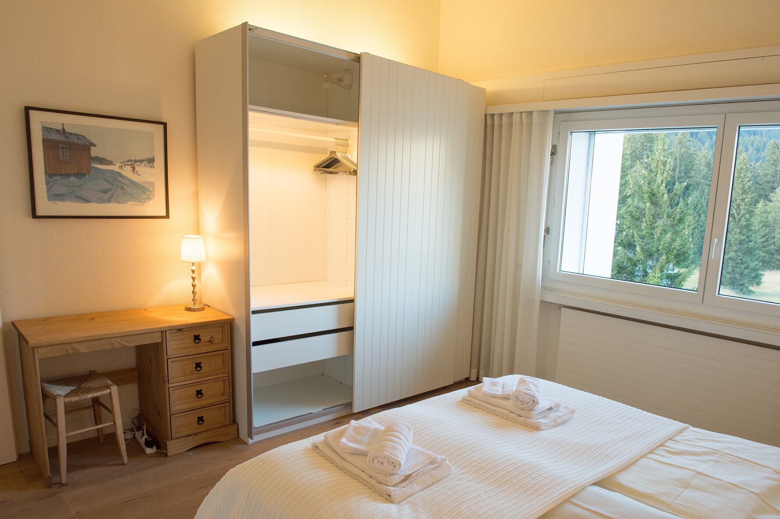 Une chambre à coucher comprenant un lit, un bureau, une armoire et une fenêtre donnant sur des arbres.
