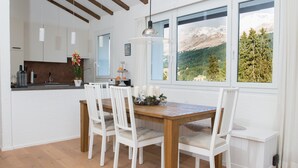A dining area with a wooden table and white chairs, a kitchen in the background, and a mountain view through large windows. - Attic apartment with pool, sauna, and fireplace (Lenzerheide/Lai)