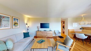 Un salon moderne avec un canapé, une table basse et des fauteuils. Un téléviseur est fixé au mur, et un meuble en bois se trouve en dessous.