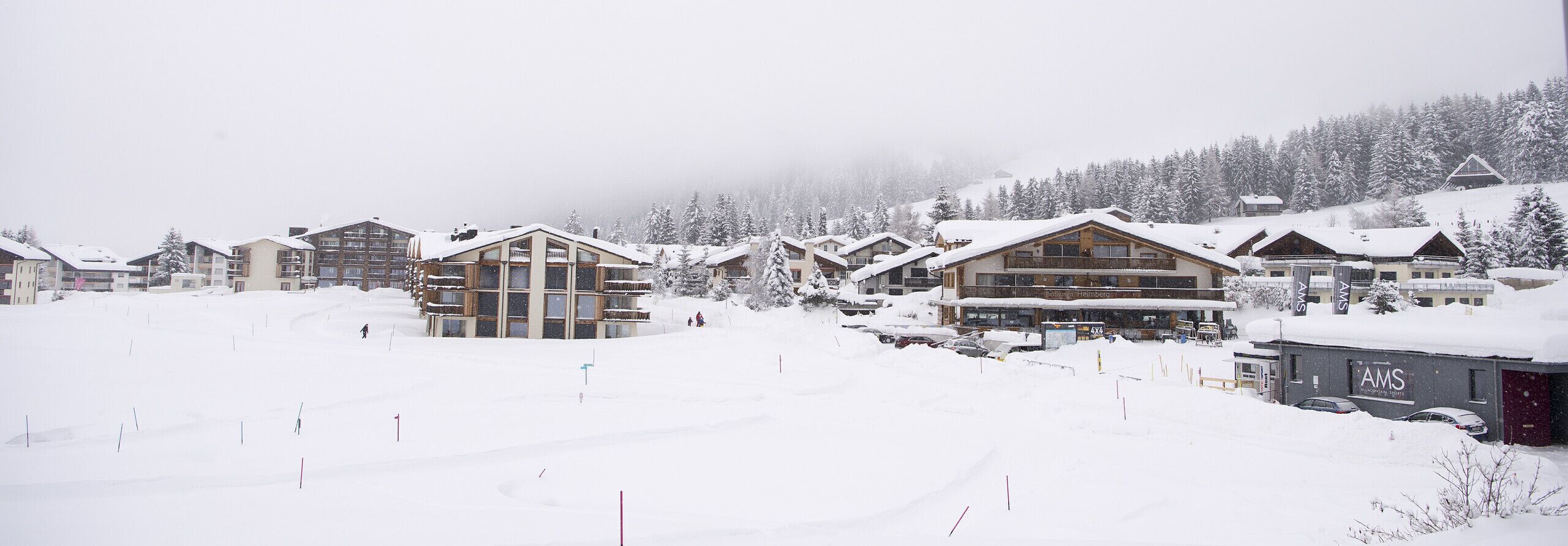 A snow-covered ski resort with chalet-style buildings, a ski lift, and a mountain backdrop.