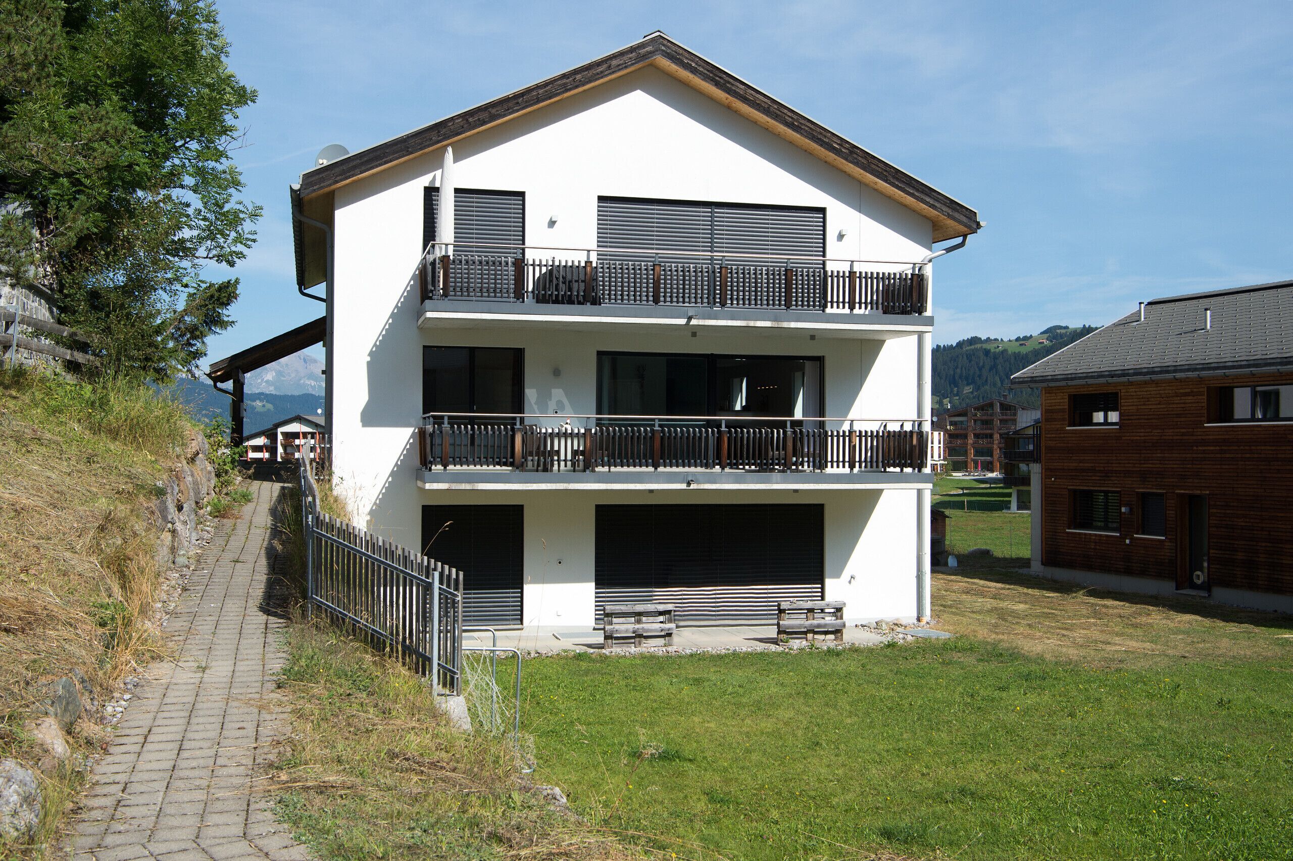 A two-story building with a balcony on the upper floor, a garage door on the ground floor, and a paved pathway leading to the entrance.